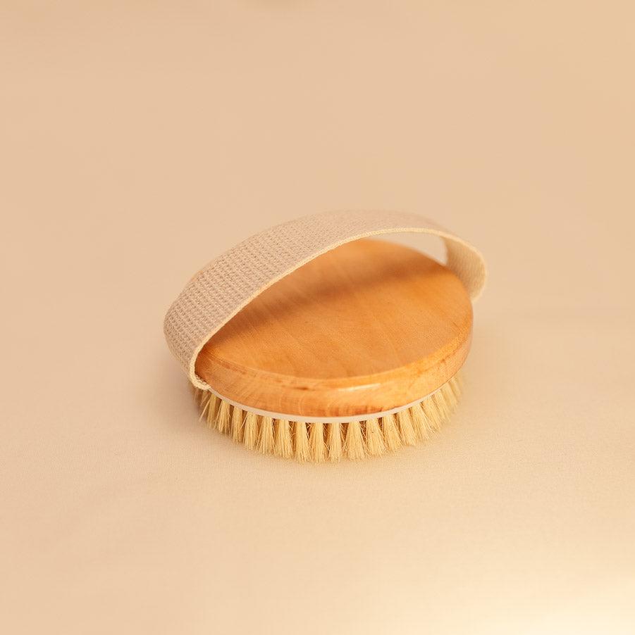 A natural bristle dry brush with a wooden handle, designed for exfoliation and lymphatic detox, placed on a clean, neutral background.